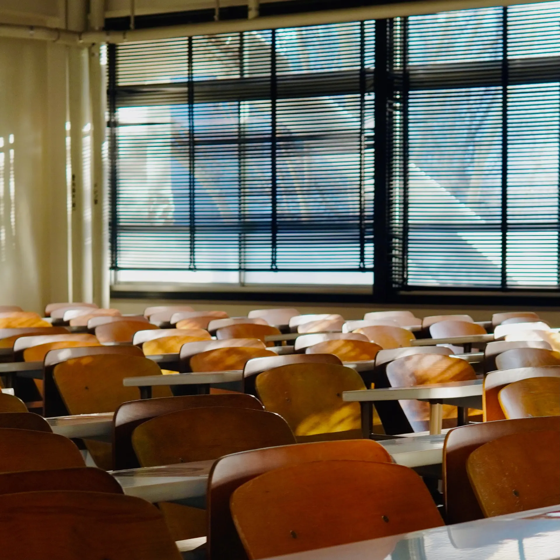 Sala de aula vazia com cadeiras de madeira iluminadas pela luz das janelas.